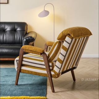A yellow-and-white striped convertible armchair sits angled on a blue area rug in a bright, modern living room. A matching striped lumbar pillow and a plush yellow headrest rest against its back, supported by dark wooden armrests and legs. In the background, a portion of a dark leather sofa and a modern floor lamp complete the interior staging.