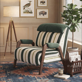A green-and-white striped convertible armchair sits on a patterned blue-and-red rug in a warmly lit, rustic room. A matching striped lumbar pillow and a dark green headrest cushion rest against its back, supported by dark wooden armrests and legs. In the background, a tripod floor lamp, framed animal art pieces, a potted indoor plant, and a wooden magazine rack complete the cozy interior staging.