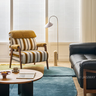A yellow-and-white striped convertible armchair sits on a colorful layered rug in a bright, modern living room. A matching striped lumbar pillow and a plush yellow headrest rest against its back, flanked by dark wooden armrests and legs. In the foreground, a round wooden coffee table holds an open book and a bowl of fruit. A modern floor lamp, a dark leather sofa, and large windows with blinds complete the cozy interior setting.