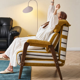 A model in a white dress sits relaxed in a yellow-and-white striped convertible armchair, stretching her arms upward. The chair features dark wooden armrests, a plush matching headrest, and a supportive lumbar pillow. She rests on a blue area rug in a sunlit living room with a black leather sofa and a modern floor lamp in the background.