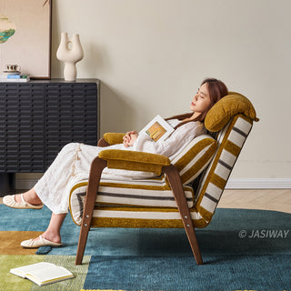 A model in a white dress rests comfortably with her eyes closed while leaning back in a yellow-and-white striped convertible armchair. The chair features dark wooden armrests, a plush matching headrest, and a supportive lumbar pillow with an open book resting on her lap. She sits atop a blue-and-green patterned area rug in a modern, warmly lit room with a dark textured cabinet in the background.