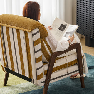 A model sits in a yellow-and-white striped convertible armchair, viewed from behind at an angle while reading an open magazine. The chair features dark wooden armrests, a plush matching headrest, and a supportive lumbar pillow. She rests atop a blue-and-green patterned area rug inside a sunlit room with sheer white curtains in the background.