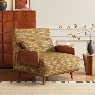 A mustard-yellow patterned fabric armchair with wooden legs and armrests sits in a warm, elegant living space. A matching lumbar pillow is neatly arranged against the backrest. In the background, a dark wood cabinet stands beside a decorative side table holding a vase with branches and a teacup.