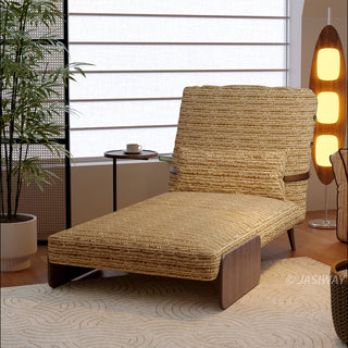 A mustard-yellow textured fabric convertible sofa is set up as a lounge chaise on a patterned rug in a sunlit room. A matching lumbar pillow is neatly placed against the upright backrest. In the background, a tall wooden floor lamp glows next to a potted plant, while a window with horizontal blinds provides soft natural light.