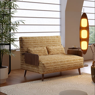 A mustard-yellow textured fabric convertible armchair sofa sits on a patterned rug in a bright, modern room. Two matching lumbar pillows rest against its slightly inclined backrest. A tall wooden floor lamp glows warmly next to the sofa, while a window with horizontal blinds provides soft natural light.