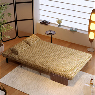 A fully flattened mustard-yellow textured fabric convertible sofa bed rests on a patterned cream rug in a bright, modern room. Two matching pillows are neatly positioned at the head of the bed. In the background, a small round side table, a tall glowing floor lamp, and a window with horizontal blinds complete the cozy setup.