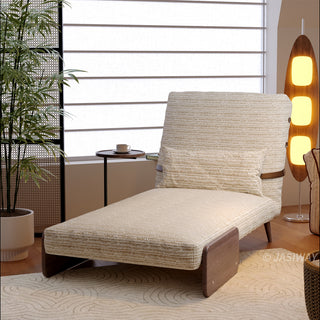 A light neutral-toned textured fabric convertible sofa is set up as a lounge chaise on a patterned rug in a bright, modern room. A matching lumbar pillow rests against its slightly tilted backrest. In the background, a tall wooden floor lamp glows warmly next to a potted plant, while a window with horizontal blinds provides soft natural light.