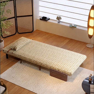 A fully flattened light neutral-toned textured fabric convertible sofa bed rests on a patterned cream rug in a bright, modern room. A matching pillow is placed neatly at the head of the bed. In the background, a tall wooden floor lamp, a woven partition screen, and a window with horizontal blinds complete the cozy setup.