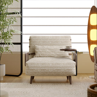 A straight-on view showcases a light neutral-toned textured fabric convertible armchair sofa set up in a modern living space. A matching lumbar bolster pillow rests against the upright backrest, and a small built-in side table holds a cup. The chair is positioned on a patterned area rug with a tall, glowing oval floor lamp and a leafy green plant nearby.