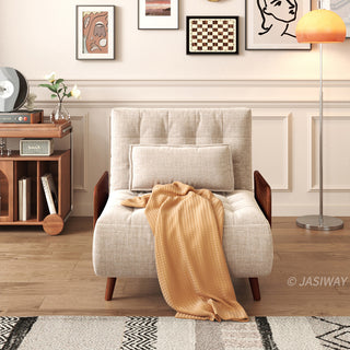 A beige linen convertible armchair sits centered against a paneled wall featuring framed artwork and a standing floor lamp. A rectangular lumbar pillow rests against its tufted back, and a warm yellow throw blanket drapes over the seat. The wooden floor is partially covered by a patterned area rug, with a media cabinet visible on the left.