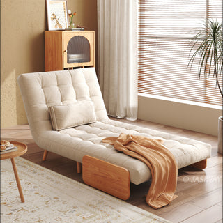 A beige linen convertible armchair is set up as an extended lounge chaise on a patterned rug in a sunlit room. A rectangular lumbar pillow rests against its tufted back, and a warm yellow throw blanket drapes across the lower seat. In the background, a wooden cabinet, white curtains, and a potted plant complete the cozy lifestyle setting.