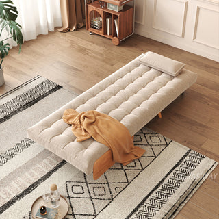 A beige linen convertible armchair is fully flattened into a sleek single bed atop a geometric patterned area rug in a sunlit room. A single rectangular pillow rests neatly at the head, while a warm yellow throw blanket is casually draped across the middle. In the background, a wooden floor, a rolling storage cart, and a potted plant complete the cozy modern living space.