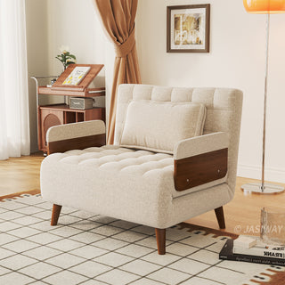 A beige linen tufted convertible armchair is set on a patterned grid rug in a sunlit living room. A single matching rectangular pillow rests against its tufted backrest, complemented by wooden armrests and tapered legs. In the background, a standing floor lamp, a framed wall artwork, and a wooden cabinet complete the cozy lifestyle setting.
