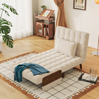 A beige linen tufted convertible armchair is set up as an extended lounge chaise on a patterned rug in a sunlit room. A rectangular pillow rests neatly against its backrest, while a blue throw blanket drapes across the lower seat. In the background, a large potted plant, a wooden storage cart, and a standing floor lamp complete the cozy lifestyle setting.