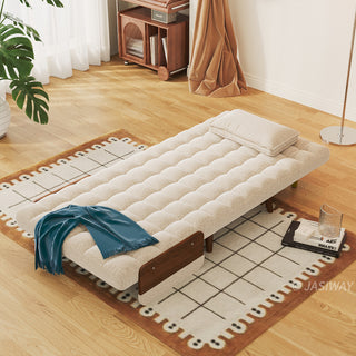 A beige linen tufted convertible armchair is fully flattened into a sleek single bed on a patterned rug in a sunlit room. A single rectangular pillow rests at the head of the bed, while a blue throw blanket is draped over the lower end. In the background, a wooden floor, a rolling storage cart, and a standing floor lamp complete the cozy modern living space.