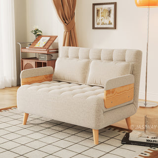 A beige linen tufted convertible loveseat sits on a patterned grid rug in a sunlit living room. Two matching pillows rest neatly against its supportive backrest, flanked by wooden armrests and tapered legs. In the background, a standing floor lamp, a framed wall artwork, and a wooden storage cart complete the cozy lifestyle setting.
