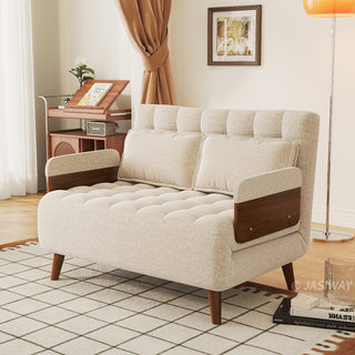 A beige linen tufted convertible loveseat sits on a patterned grid rug in a sunlit living room. Two matching pillows rest neatly against its supportive backrest, flanked by wooden armrests and tapered legs. In the background, a standing floor lamp, a framed wall artwork, and a wooden storage cart complete the cozy lifestyle setting.
