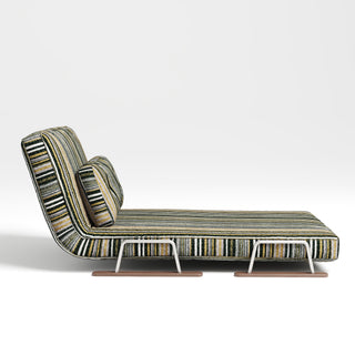 A convertible lounge chair is shown from a side profile against a plain, neutral background. The piece is fully extended in a reclined or lounger position with an angled backrest and an extra pillow. It is upholstered in a textured fabric featuring multicolored horizontal stripes. The frame is supported by U-shaped metal legs attached to flat wooden base rails.