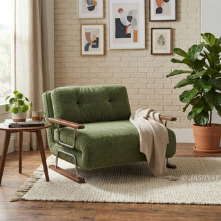 A modern armchair upholstered in textured green fabric sits on a light-colored, chunky-knit area rug over a wood floor. The chair features button-tufted back cushions, wooden armrests, a sleek sled base, and a draped beige throw blanket. In the background, a white brick accent wall is decorated with a gallery wall of framed abstract art, complemented by a small side table and a large potted fiddle-leaf fig.