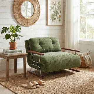 A modern armchair upholstered in a textured green fabric sits on a woven jute rug over a light wooden floor. The chair features button-tufted cushions, dark wooden armrests, and a sleek sled base. To the left, a small wooden side table holds a potted plant and books, while a white-paneled wall decorated with a round woven mirror and botanical print creates a warm, natural aesthetic.