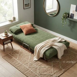 A modern convertible daybed upholstered in textured green fabric rests fully extended on a patterned jute rug over a light wooden floor. The daybed is styled with a plush rust-orange accent pillow and a draped cream-knit throw blanket. A small mid-century modern wooden side table sits beside it, while a dark green accent wall adorned with a round mirror and a hanging plant completes the room.