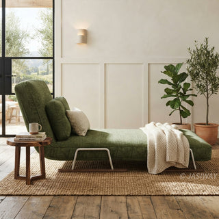 A modern chaise lounge upholstered in textured green fabric is positioned in a tranquil, sunlit room on a woven jute rug over a wooden floor. The lounge features an adjustable backrest, a matching lumbar pillow, and a draped cream knit throw blanket. A small live-edge wooden side table sits at the head, while potted greenery and a neutral paneled wall complete the serene setting.