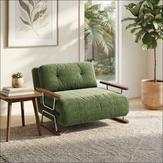 A modern armchair upholstered in textured green fabric rests on a light-colored woven rug over a wooden floor. The chair features button-tufted back cushions, wooden armrests, and a unique sled base. In a sunlit room, a small wooden side table holds a potted succulent, while a large potted fiddle-leaf fig tree and a botanical wall art print complete the natural decor.