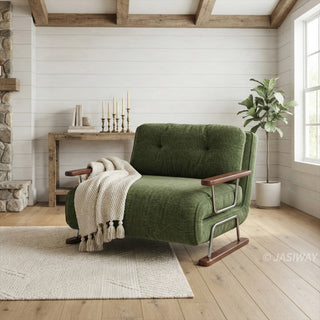 A modern armchair upholstered in textured green fabric rests on a light-colored area rug over a hardwood floor. The chair features button-tufted back cushions, wooden armrests, a sleek sled base, and a draped cream-knit throw blanket with tassels. In the background, a rustic living room with a white-paneled wall, exposed wooden ceiling beams, a stone fireplace, and a potted plant creates a cozy, inviting atmosphere.