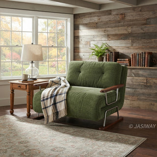 A modern armchair upholstered in textured green fabric is positioned in a rustic room featuring a reclaimed wood accent wall and hardwood flooring. The chair features button-tufted back cushions, wooden armrests, a sleek sled base, and a draped plaid throw blanket. Beside it, a wooden side table holds a table lamp, while a large window lets in natural light.