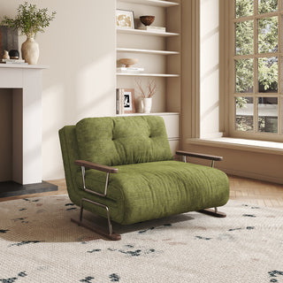 A modern living room interior features a convertible armchair sofa with a distinctive green textured fabric upholstery and tufted backrest. The sofa bed is positioned centrally on a large, patterned area rug, showcasing its dark wood armrests and chrome and dark wood frame. In the background, a built-in white bookshelf filled with various decor items and books stands beside a large window that overlooks lush greenery.