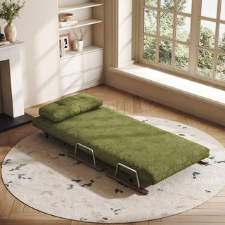 A fully reclined green textured fabric convertible sofa bed rests on a round patterned rug in a bright, sunlit room. A supportive matching bolster pillow is placed at the head of the daybed. In the background, a large multi-pane window floods the wooden-floored space with natural light.