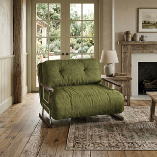 A rustic, sunlit living room features a green textured fabric armchair sofa with dark wood armrests and metal legs, placed on a patterned rug. In the background, a wooden fireplace with a stone surround sits beside a side table with a lamp, while double glass doors look out onto an outdoor green garden view.
