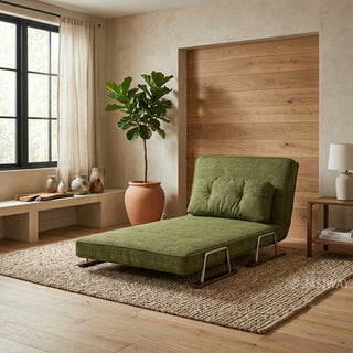 A green textured fabric convertible sofa is set up as a lounge chair on a textured jute rug in a warm, minimalist room. A matching bolster pillow rests against its slightly inclined backrest. In the background, a large potted plant stands near a window with sheer curtains, alongside a wooden accent wall panel and a small side table with a lamp.