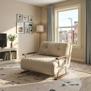 A cozy, modern living room features a beige fabric armchair with dark wooden armrests and metal legs placed centrally on a round patterned rug. In the background, a window framed by grey curtains looks out onto city buildings and power lines. A media console with a TV, framed wall art, and a floor lamp complete the warm, sunlit interior.