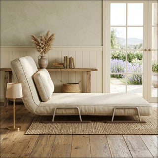 A profile view of a modern beige textured fabric convertible sofa is set up as a chaise lounge on a jute rug. The adjustable backrest features an attached lumbar pillow and rests on sleek metal and wood sled-style legs. In the background, a rustic wooden console table, a small brass desk lamp, and double glass doors leading to a blooming garden complete the warm, sunlit country-style room.