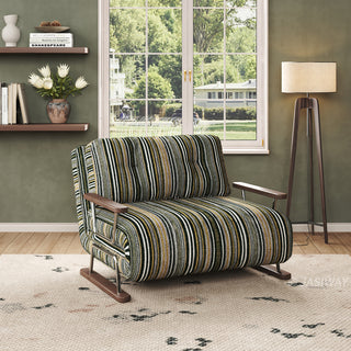 A modern armchair upholstered in a dark striped fabric with green, black, and beige tones sits on a light-colored patterned area rug over a wooden floor. The chair features button-tufted back cushions, wooden armrests, and a sleek sled base. In the background, an olive-green wall with floating shelves, a vase of white flowers, a large window with a garden view, and a standing floor lamp complete the tranquil, natural aesthetic.