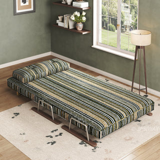 A modern convertible daybed upholstered in dark striped fabric with green, black, and beige tones is fully extended on a light-colored patterned area rug over a wooden floor. The daybed features a supportive tufted back cushion positioned at the head and a sleek sled base design. In the background, an olive-green wall with floating shelves, wall art, a window with natural light, and a standing floor lamp complete the tranquil room setup.
