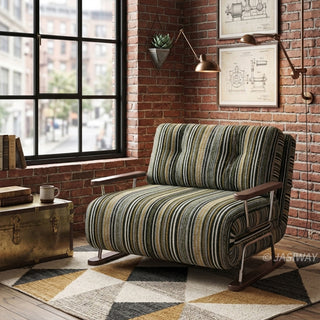 A modern armchair upholstered in dark striped fabric with green, black, and beige tones sits on a geometric patterned area rug over a wooden floor. The chair features button-tufted back cushions, wooden armrests, and a sleek sled base. In the background, an industrial loft setting with exposed red brick walls, a vintage metal chest as a side table, framed architectural blueprints, and a large grid window completes the rustic urban aesthetic.