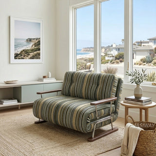 A modern armchair upholstered in dark striped fabric with green, black, and beige tones is positioned on a neutral jute area rug over a wooden floor. The chair features button-tufted back cushions, wooden armrests, and a sleek sled base. In the background, a coastal-inspired living room with large windows overlooking a scenic beach view, coastal wall art, and a small side table with a potted plant completes the bright, airy aesthetic.