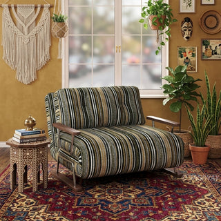 A modern armchair upholstered in dark striped fabric with green, black, and beige tones sits on a vibrant, patterned bohemian area rug over a wooden floor. The chair features button-tufted back cushions, wooden armrests, and a sleek sled base. In the background, an eclectic bohemian room with a macrame wall hanging, carved side table, potted plants, and wall decor completes the warm, artistic aesthetic.