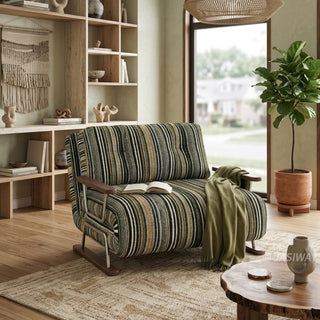 A modern armchair upholstered in dark striped fabric with green, black, and beige tones sits on a neutral textured area rug over a wooden floor, draped with a cozy olive-green throw blanket and an open book. The chair features button-tufted back cushions, wooden armrests, and a sleek sled base. In the background, a warm, organic living space with built-in wooden shelving, a woven macrame wall hanging, a large window, and a potted fiddle-leaf fig tree complete the inviting decor.
