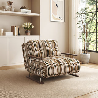 A modern armchair upholstered in a warm brown, black, and beige striped fabric sits on a light-colored woven area rug over a wooden floor. The chair features button-tufted back cushions, wooden armrests, and a sleek sled base. In the background, a sunlit room with built-in shelving, minimalist wall art, and a potted plant creates a cozy, modern bohemian atmosphere.
