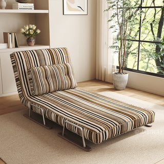 A modern chaise lounge upholstered in warm brown, black, and beige striped fabric is set up in a sunlit room on an area rug over a wooden floor. The lounge features an adjustable backrest, a matching lumbar pillow, and a sleek sled base. In the background, built-in shelving, minimalist wall art, and a large window with natural light complete the cozy setup.