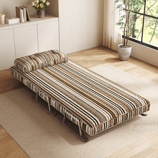 A modern convertible daybed upholstered in warm brown, black, and beige striped fabric is fully extended on a light-colored area rug over a wooden floor. The daybed features a supportive back cushion attached at the head and a sleek sled base design. In the background, a bright, minimalist room with built-in cabinetry, books, and a potted plant by a large window completes the clean, modern look.