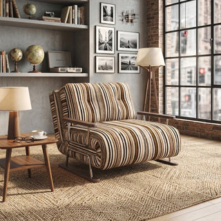 A modern armchair upholstered in a warm brown, black, and beige striped fabric sits on a patterned jute rug over a wooden floor. The chair features button-tufted back cushions, wooden armrests, and a sleek sled base. In the background, an industrial loft setting with a concrete wall, a gallery wall of black-and-white prints, a side table with a lamp, and a large grid window completes the urban aesthetic.