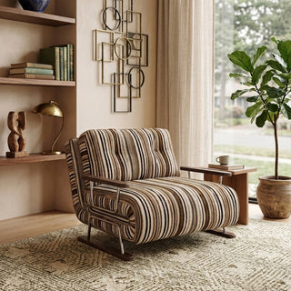 A modern armchair upholstered in warm brown, black, and beige striped fabric sits on a patterned neutral area rug over a wooden floor. The chair features button-tufted back cushions, wooden armrests, and a sleek sled base. In the background, a warm, contemporary living space with geometric wall art, built-in wooden shelving, and a potted fiddle-leaf fig tree by the window completes the inviting decor.