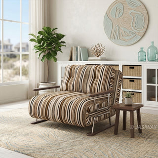 A modern armchair upholstered in warm brown, black, and beige striped fabric sits on a light-colored patterned area rug over a wooden floor. The chair features button-tufted back cushions, wooden armrests, and a sleek sled base. In the background, a coastal-inspired living room with a white storage unit, woven baskets, a wall hanging, and natural light from a large window completes the bright, airy aesthetic.
