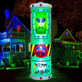 Giant Halloween inflatable alien in a green containment tube pod yard decoration with glowing LED lights, hazard markings, and "Happy Halloween" lettering, displayed on a residential front lawn at night.