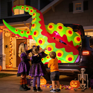 Three children in Halloween costumes trick-or-treating at a decorated car trunk featuring a giant green and red monster tentacle inflatable yard decoration, surrounded by pumpkins and a skeleton in a suburban driveway at night.