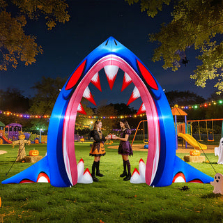 Giant inflatable shark mouth archway yard and holiday decoration with glowing lights and sharp teeth, displayed in a park at night with children in Halloween costumes standing underneath.